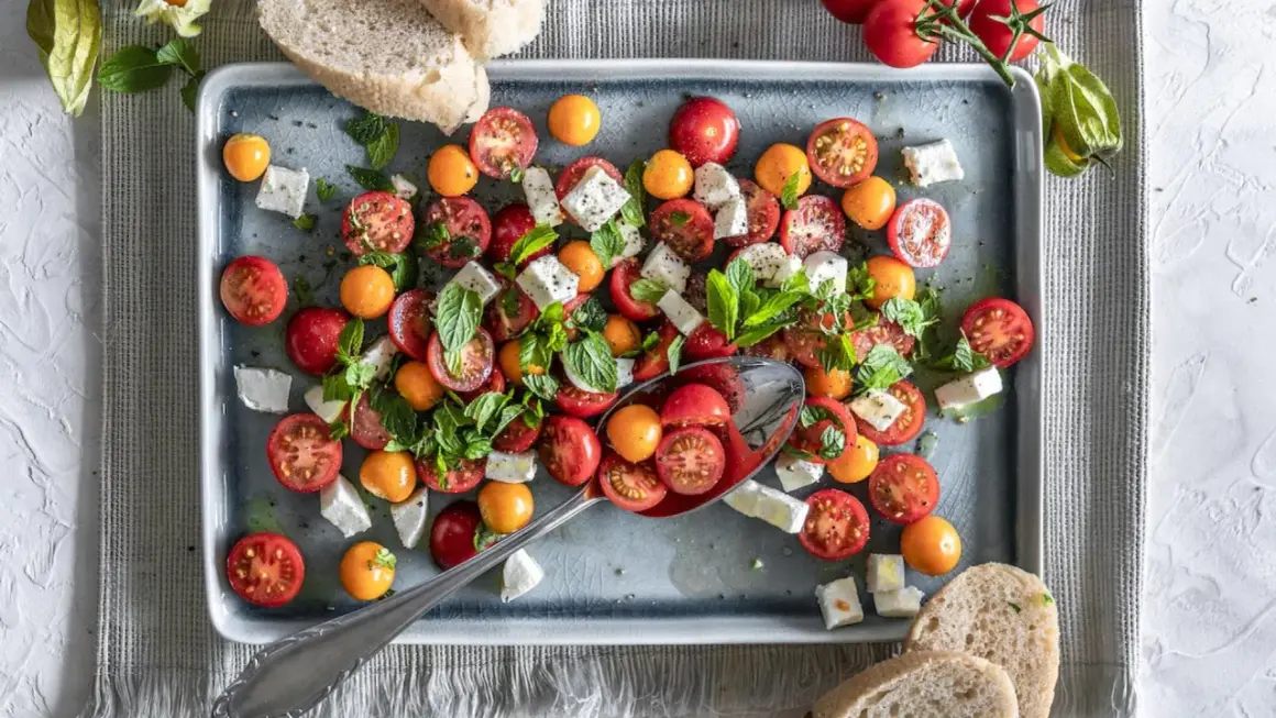 Frischer Tomate Mozzarella Salat mit bunten Kirschtomaten, Feta und Minze, serviert auf einem grauen Teller mit Brot.