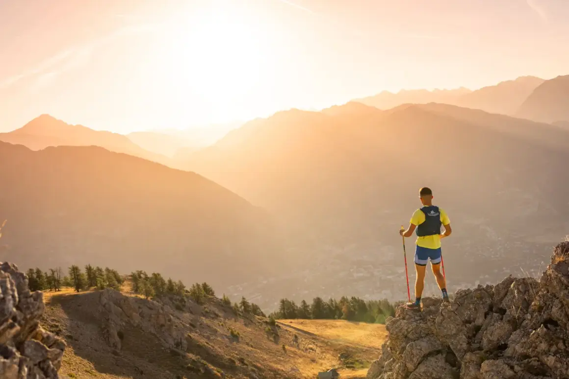 Un coureur de trail, bâtons en main, admire le paysage montagneux au lever du soleil.