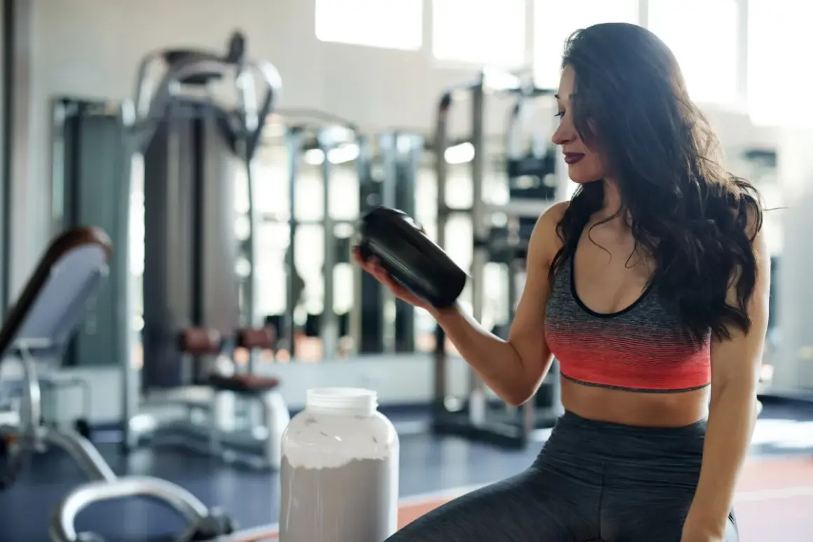 Frau im Fitnessstudio hält einen Shaker, daneben ein großer Behälter mit Pulver. Perfekt für den **proteinshake vor oder nach dem training**.