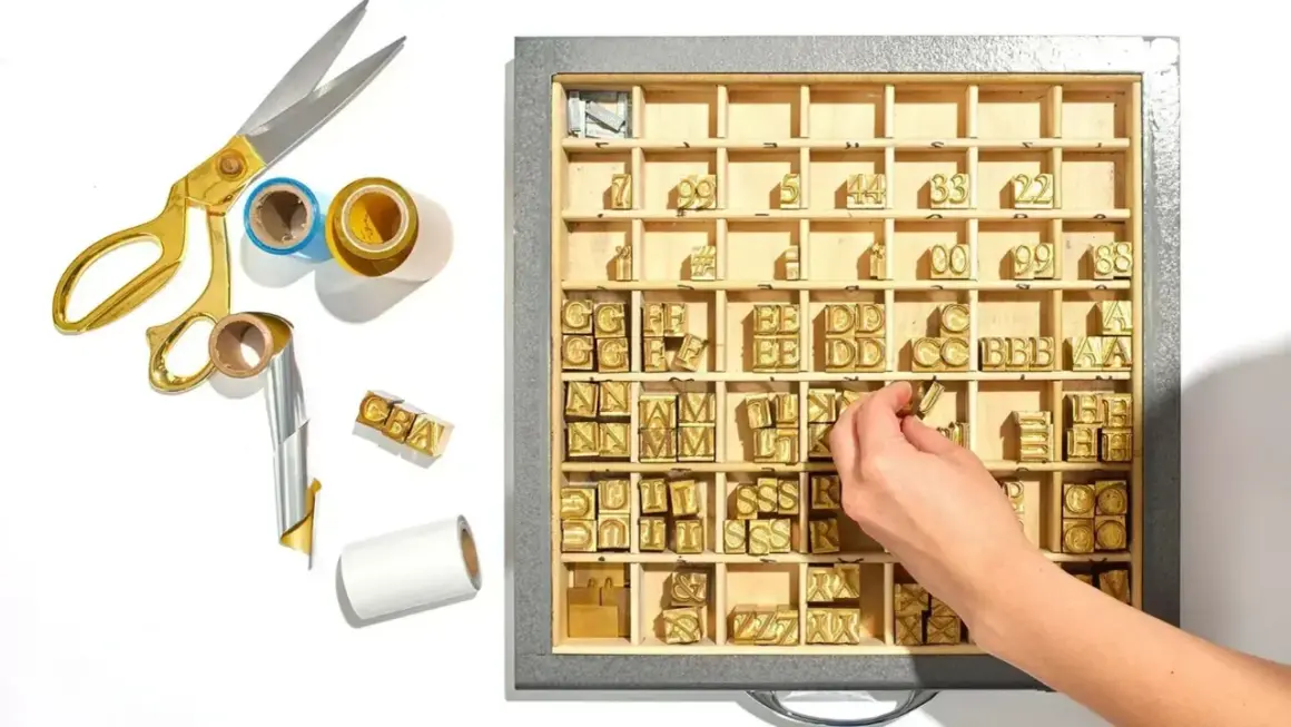 A hand selects a letter stamp from a tray of vintage letterpress type, part of a collection for creating a monogram.