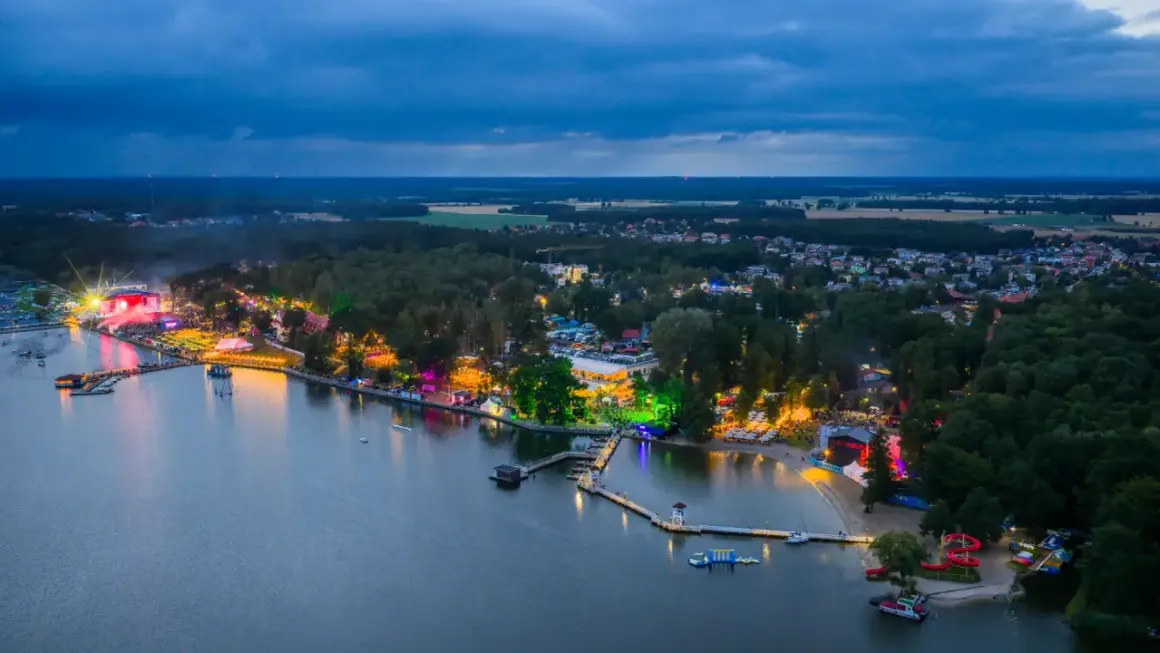 Nocny widok na jezioro i rozświetlone tereny imprezowe. Widać scenę, tłumy ludzi i kolorowe światła, które tworzą atmosferę sława festiwal.
