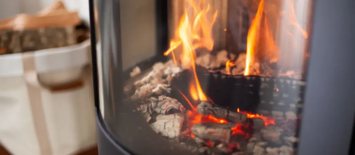 Glühende Kohlen und Flammen in einem modernen Kaminofen. Links daneben ein Korb mit Holzscheiten.