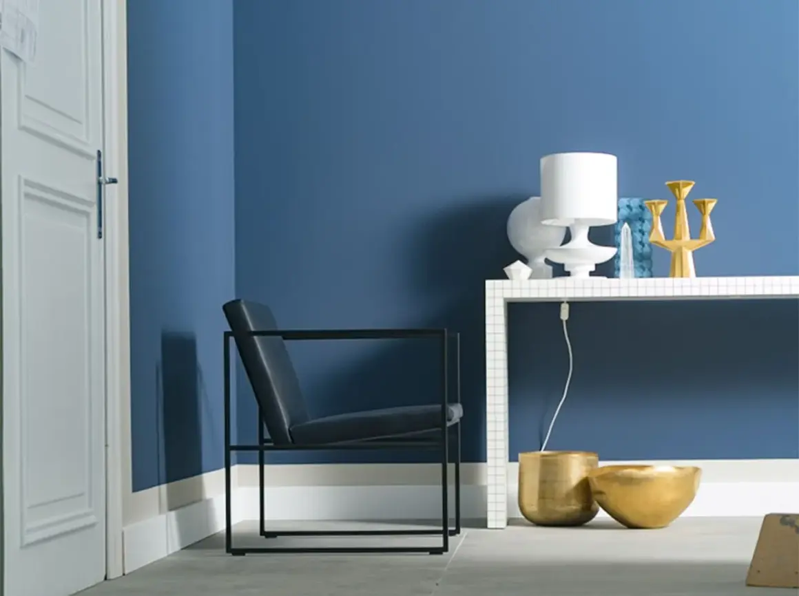 Moderne Ecke mit schwarzem Sessel, weißer Tür und blau gestrichener Wand. Die Wirkung der Wandfarbe wird durch goldene Vasen und eine Lampe auf einem Tisch betont.