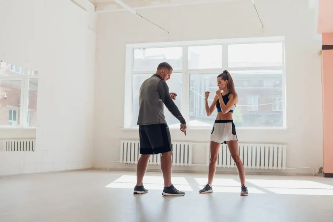A trainer instructs a woman on her right hand boxing stance in a bright, airy gym.