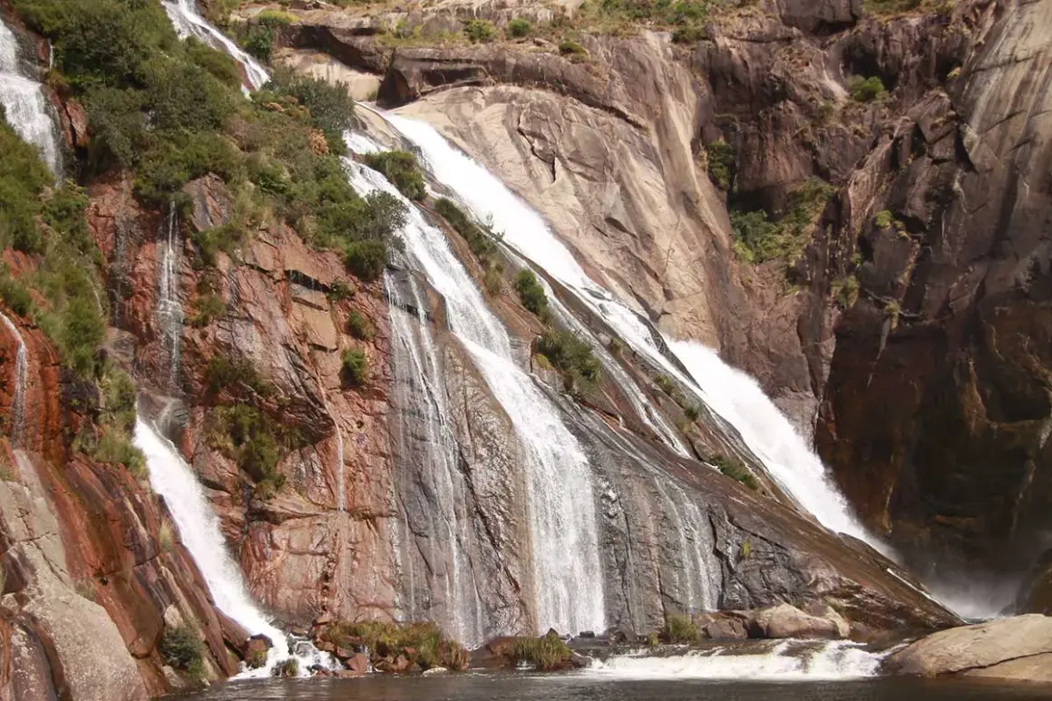Cascada de agua blanca cayendo por rocas rojizas y grises, rodeada de vegetación. Un espectáculo natural de las fervenzas de Galicia.