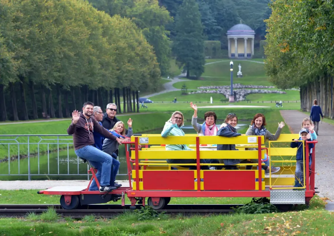 Fröhliche Menschen genießen eine Draisinenfahrt NRW durch eine Parklandschaft mit einem historischen Gebäude im Hintergrund.