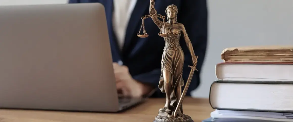 Justitia-Statue neben Laptop und Büchern. Ein Anwalt prüft Unterlagen, vielleicht wegen eines Widerspruchs Pflegegrad.