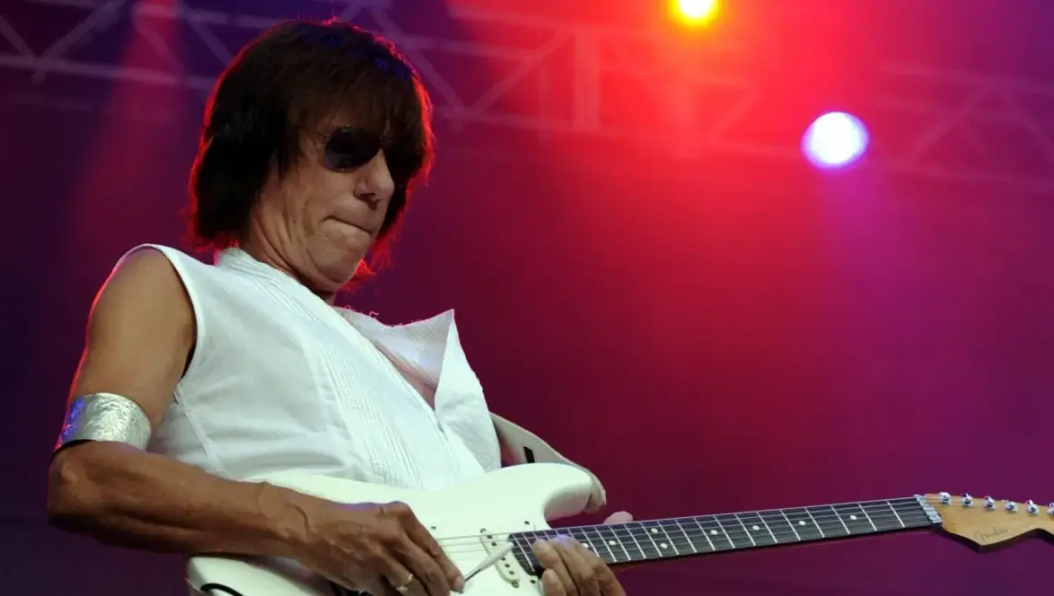 Jeff Beck, con occhiali da sole e una maglietta bianca, suona la chitarra elettrica con il suo inconfondibile stile. La sua musica, un'eredità del **the Jeff Beck group**, risuona sotto le luci rosse del palco.