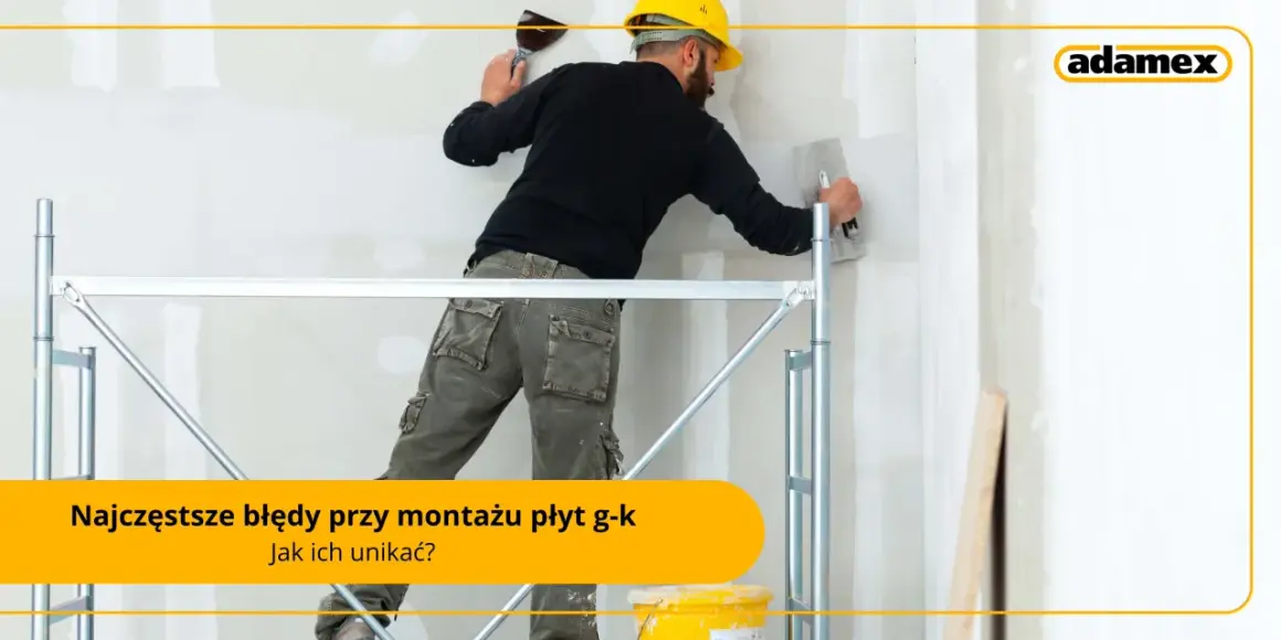 Fachowiec w żółtym kasku i roboczych spodniach na rusztowaniu wygładza ścianę z regipsu.