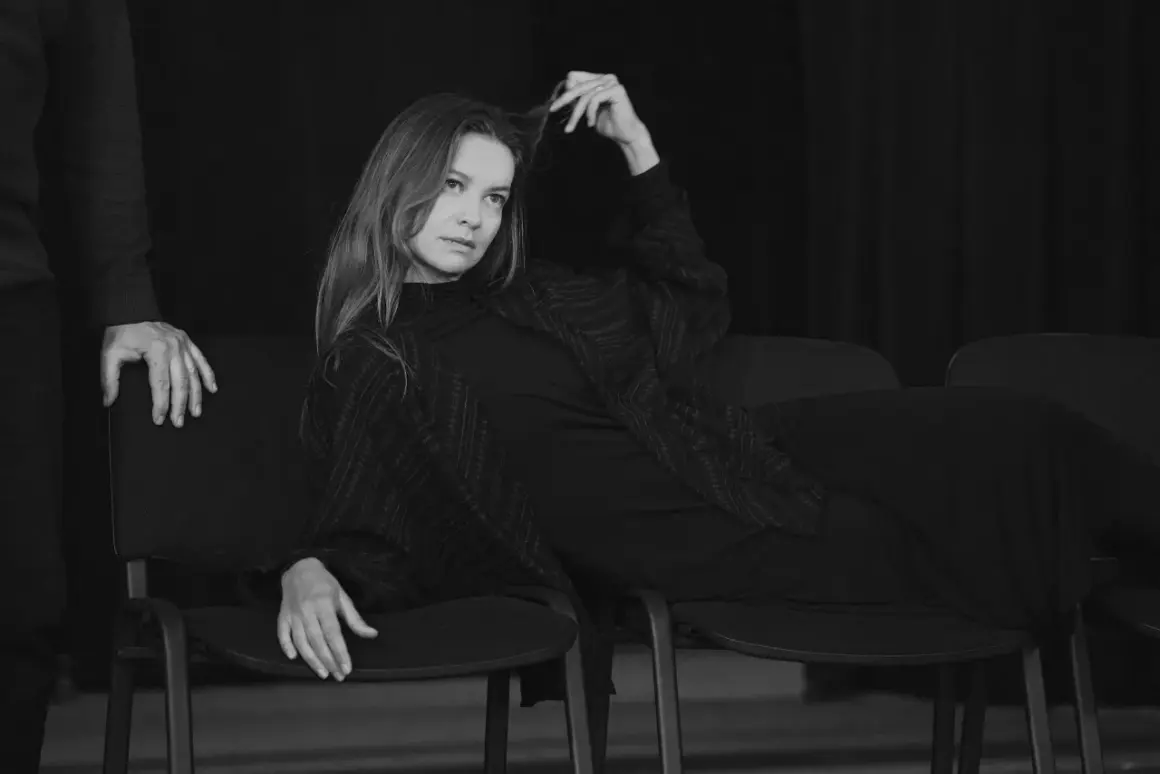 Kobieta siedzi na krześle, bawiąc się włosami. Jej spojrzenie jest nieobecne, jakby była duchem, upiorem z innego świata.