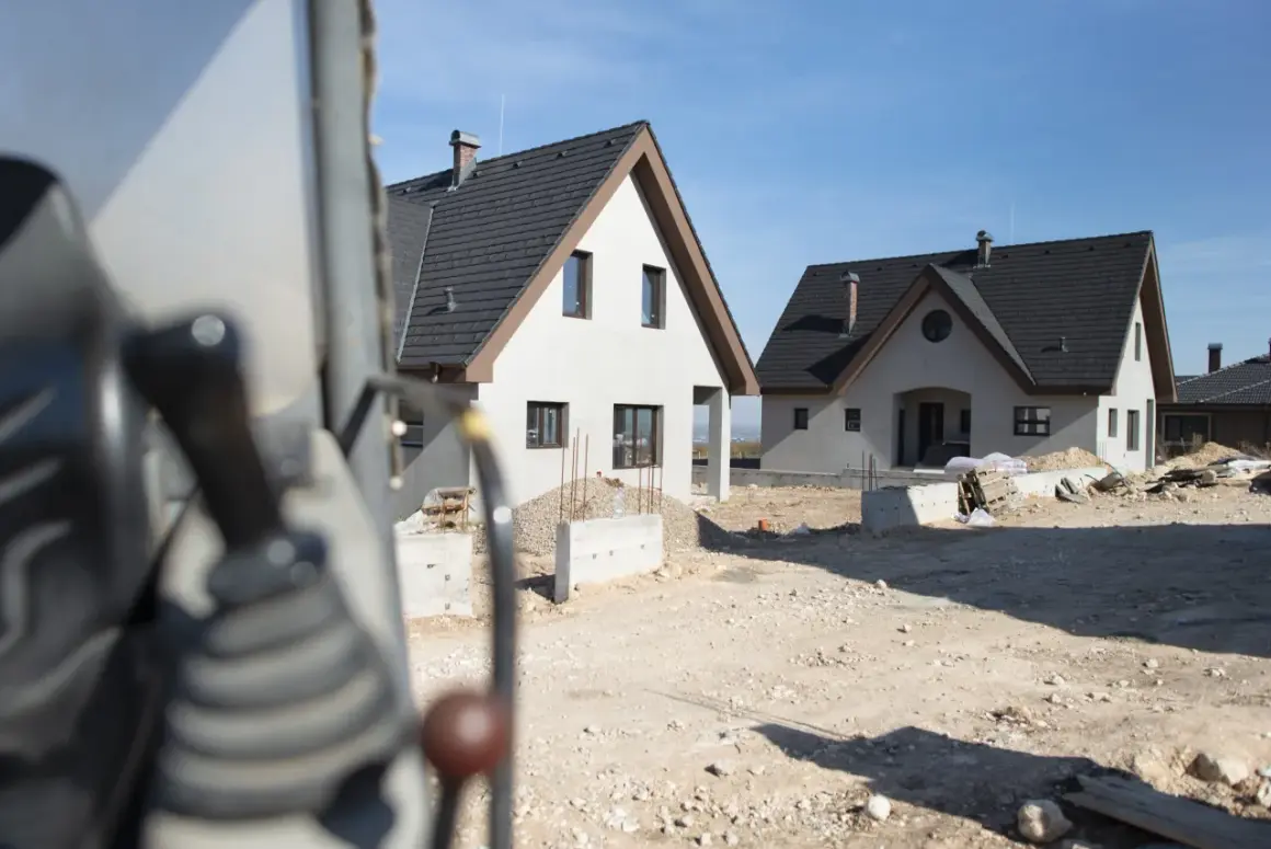 Budowa dwóch domów gospodarczych w słoneczny dzień. Widać fragment maszyny budowlanej na pierwszym planie.