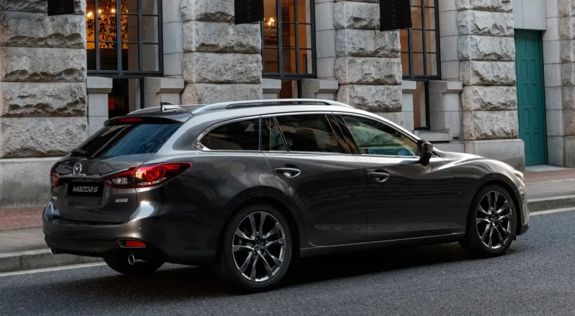 Eleganckie, ciemnoszare kombi Mazda 6, największe kombi na rynku, parkuje przed kamienną fasadą.