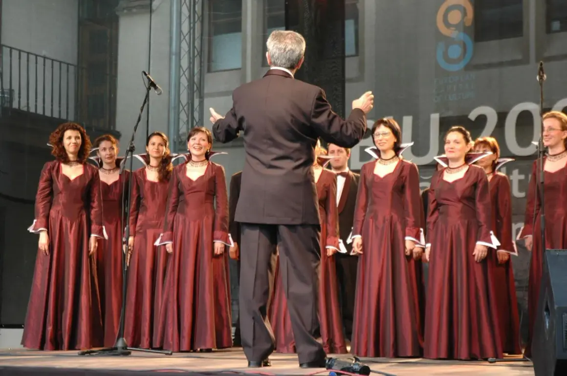 Chór kobiecy w bordowych sukniach, dyrygowany przez mężczyznę w garniturze. W tle logo "European Capital of Culture".