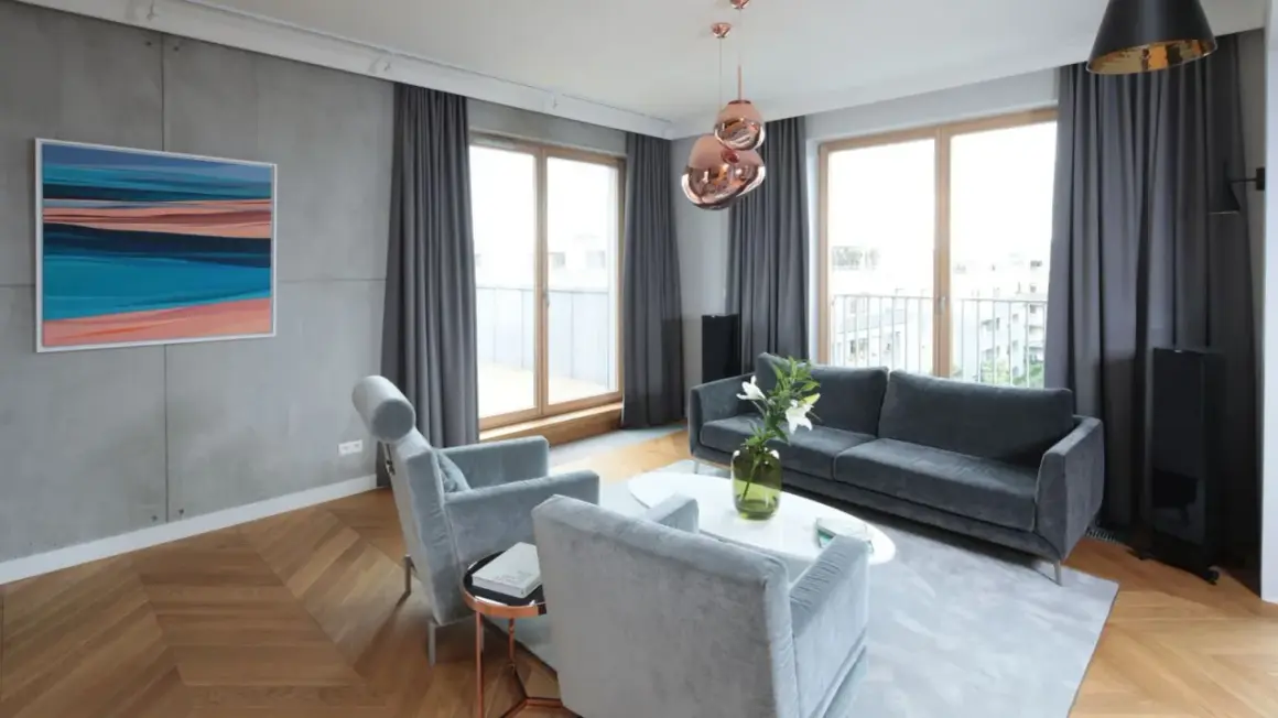 Nowoczesny salon z betonową ścianą, drewnianą podłogą w jodełkę, szarymi meblami i miedzianymi lampami. Lamele i beton tworzą industrialny klimat.