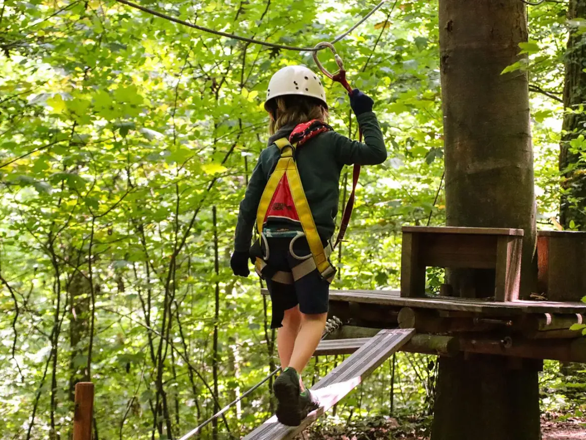 Kind im Hochseilgarten K1 balanciert auf einem schmalen Steg, gesichert durch Klettergurt und Helm.
