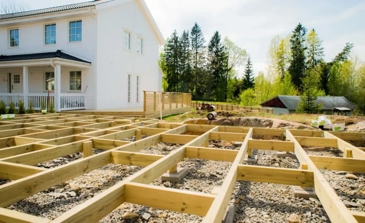 Grundläggande stomme för att bygga veranda framför ett vitt hus. Träbjälkar ligger på grus och stenar, redo för nästa steg.