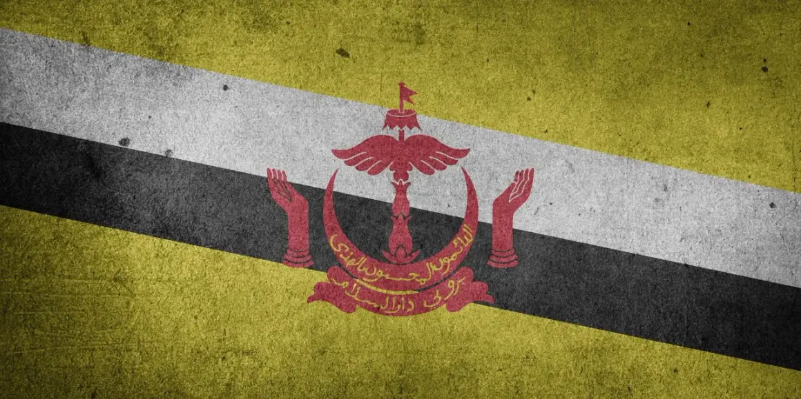 Flaga Brunei z godłem w kolorze czerwonym, na tle ukośnych pasów: żółtego, białego i czarnego.