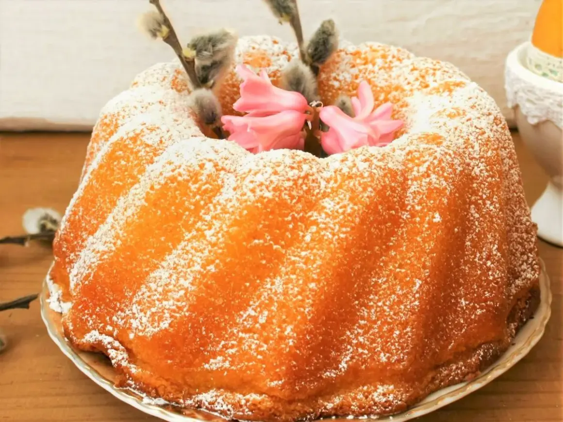Puszysta babka jogurtowa, oprószona cukrem pudrem, ozdobiona bazieczkami i różowymi kwiatami. Idealna na Wielkanoc.