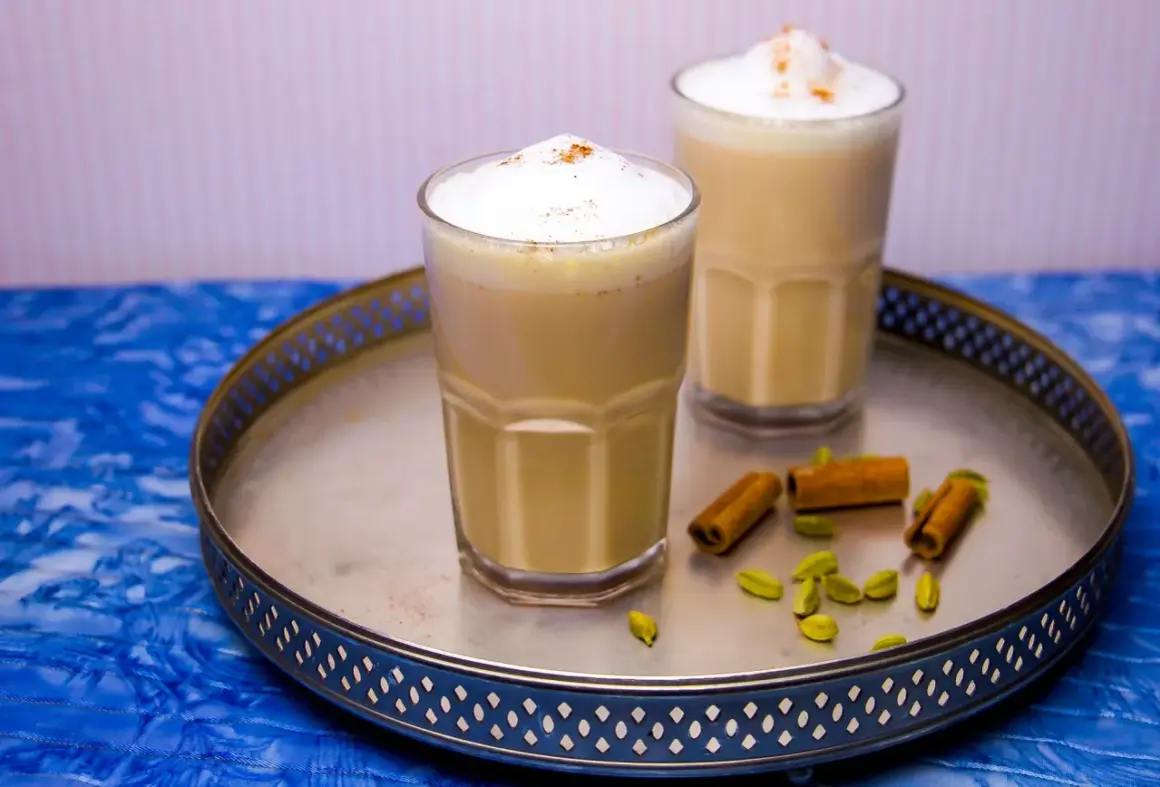 Två glas med krämig chai latte, toppad med skum och kanel. Kardemumma och kanelstänger på brickan ger en hint om detta chai latte recept.