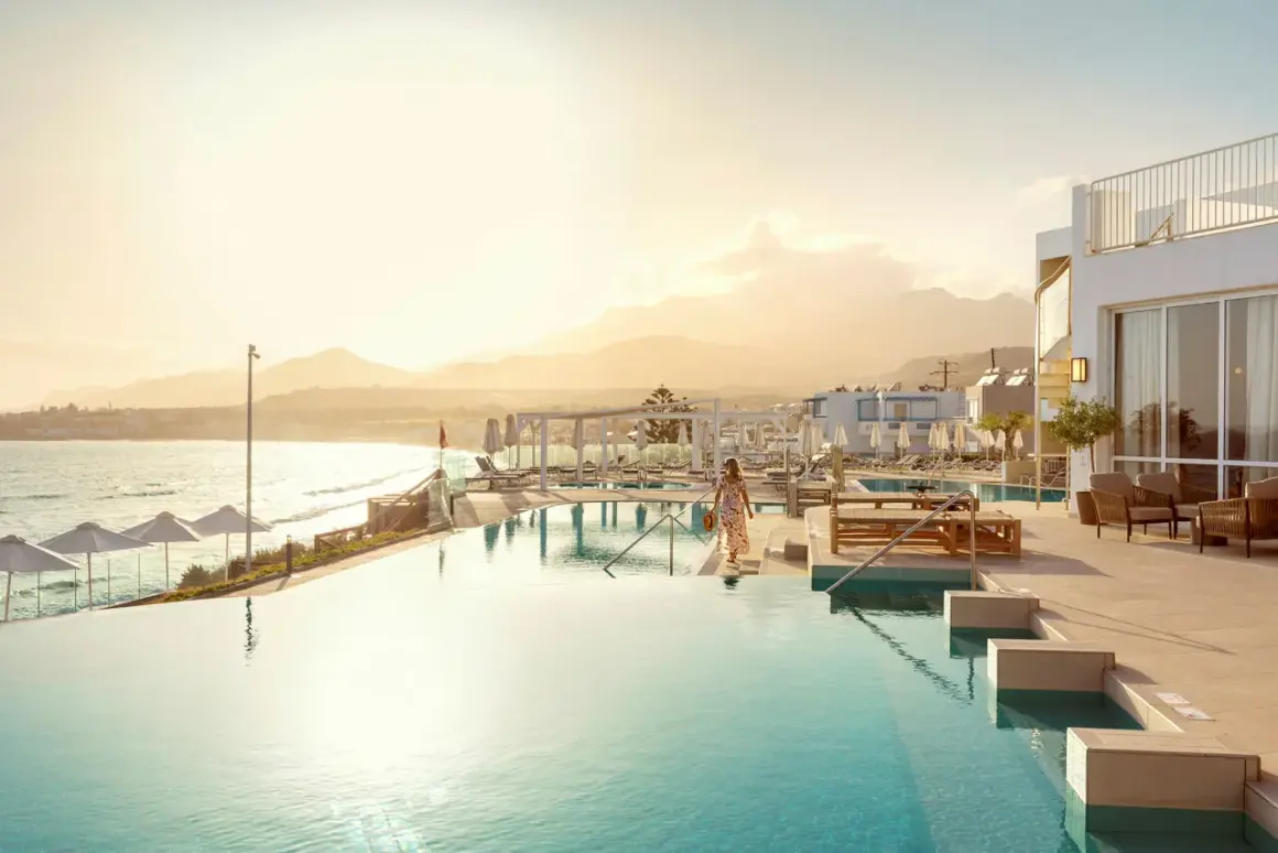 En kvinna i en blommig klänning går mot en infinitypool vid havet på Kreta, Makrigialos. Solnedgången färgar himlen gyllene.