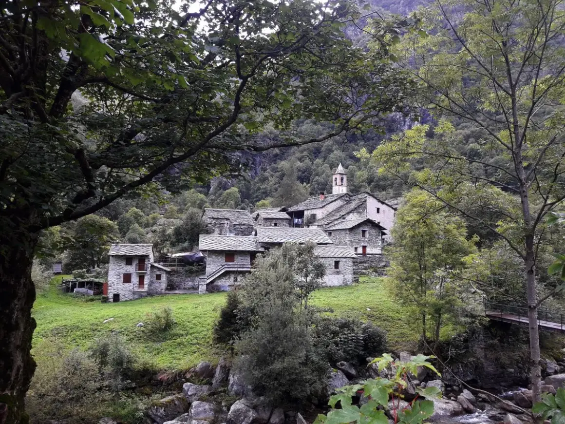 Borgo in pietra con campanile, immerso nel verde della Val Chiuse lla. Un ponte attraversa il torrente.