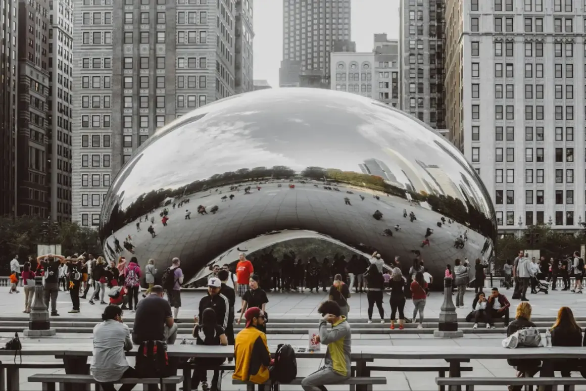 A crowd gathers around Cloud Gate, a reflective public art installation in Chicago, mirroring the city's skyscrapers and sky.