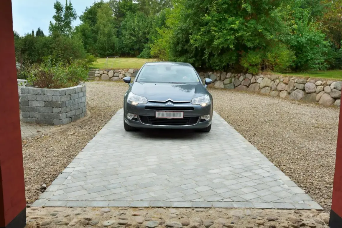 Grå bil parkerad på en vacker betong uppfart. Omgiven av grönskande träd och en stenmur.