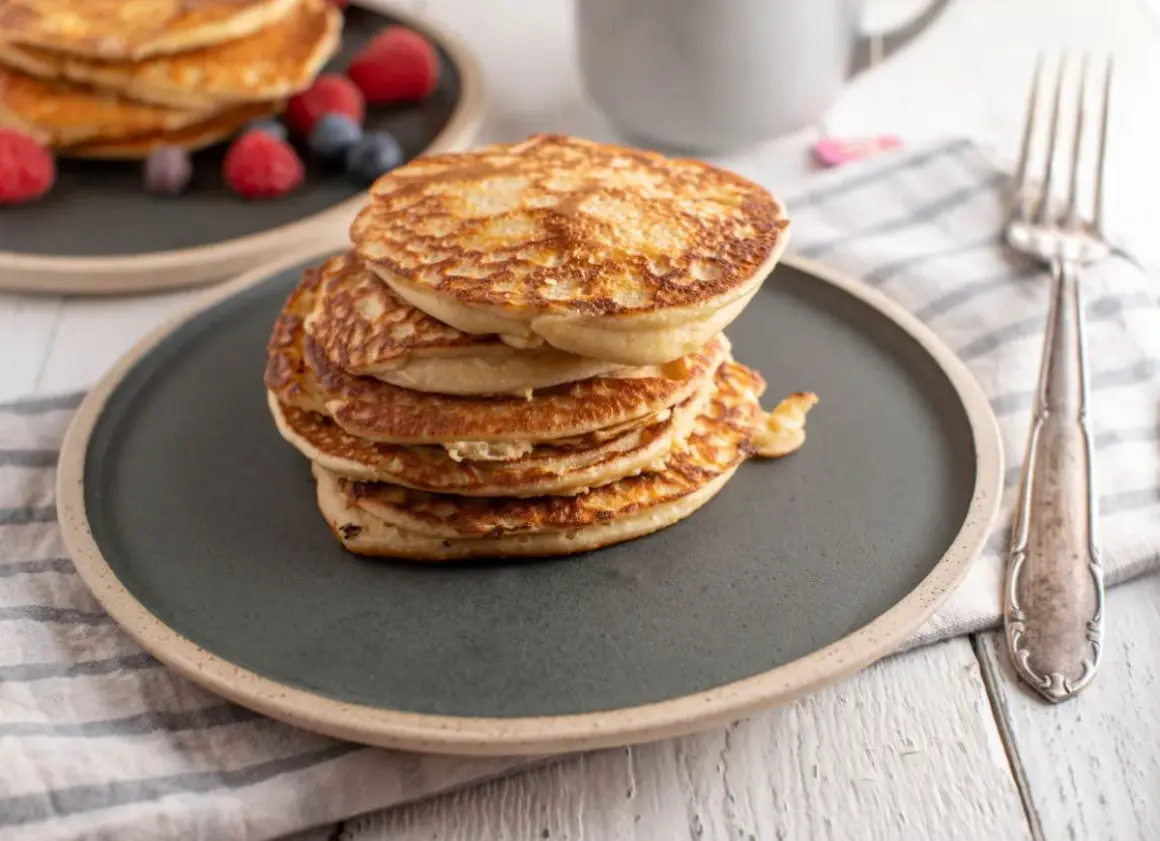Stos puszystych fit pancakes z odżywki białkowej, ozdobiony malinami i borówkami, czeka na widelcu.
