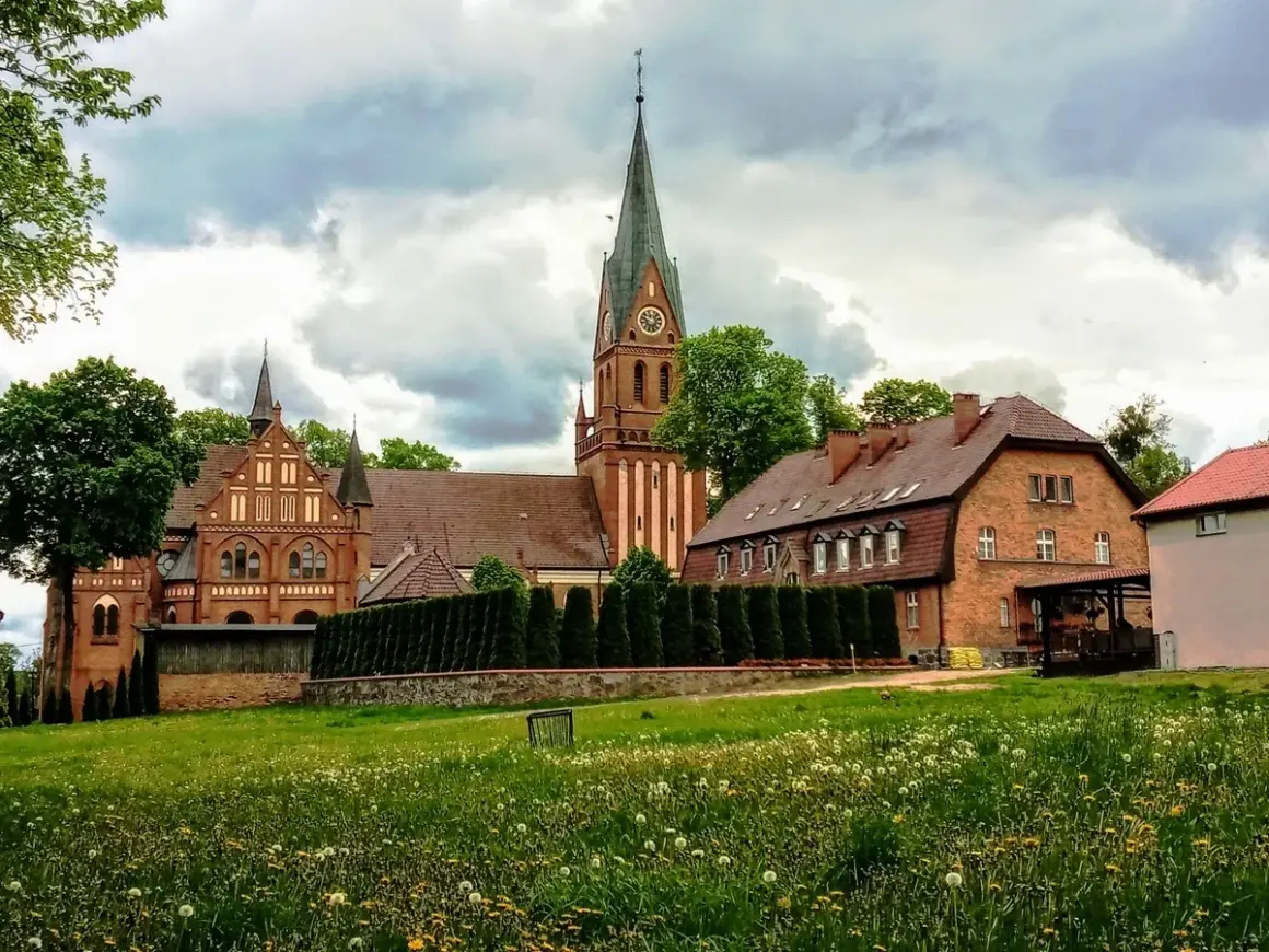 Majestatyczne sanktuarium w Gietrzwałdzie, z ceglaną architekturą i strzelistą wieżą, góruje nad zieloną łąką pełną kwiatów.
