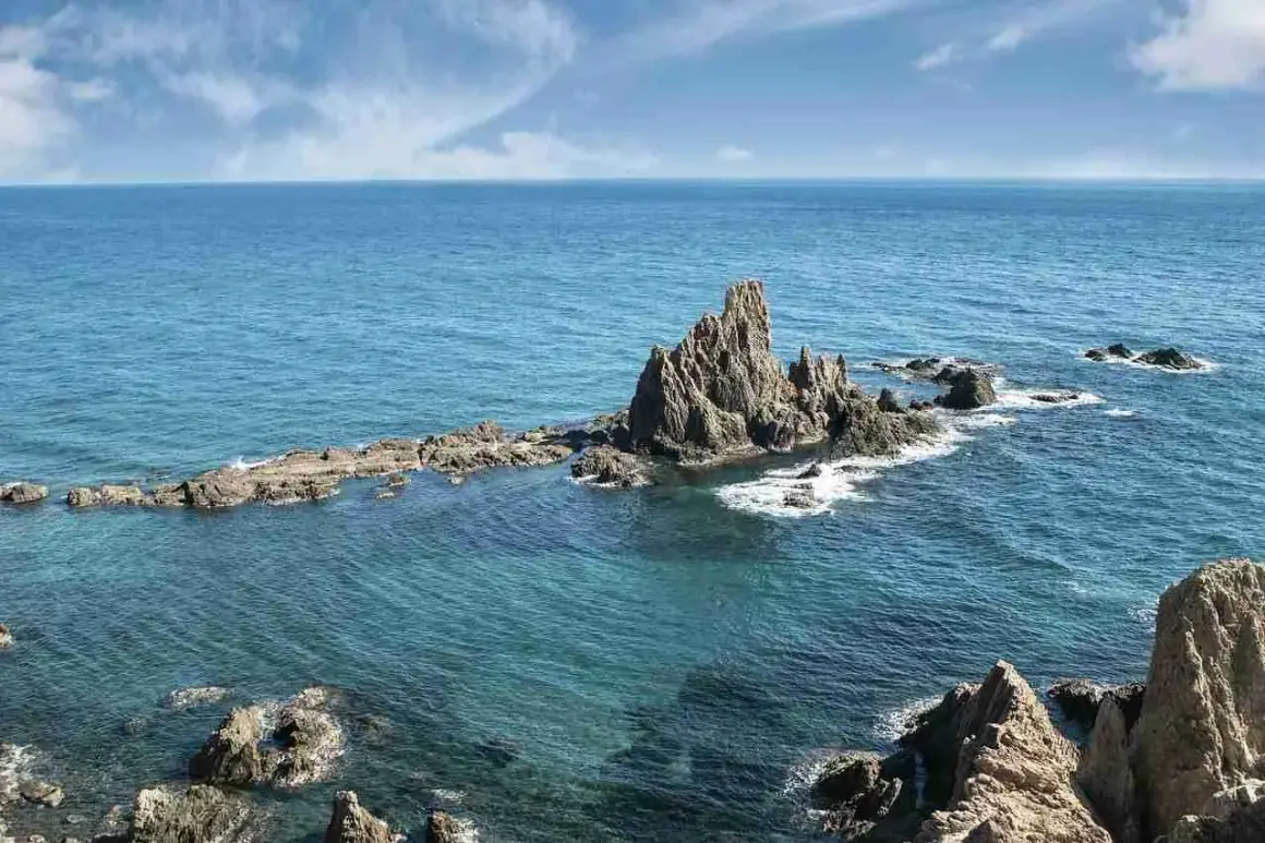 Formaciones rocosas emergen del mar turquesa en la **sierra de Cabo de Gata**. El sol ilumina las aguas cristalinas y el cielo azul con nubes blancas.