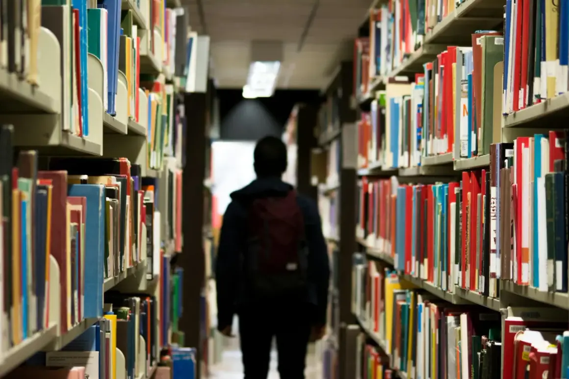 Ein Schüler mit Rucksack geht durch eine Bibliothek. Vielleicht sucht er nach Büchern, die ihm zeigen, dass das Gymnasium viel schwerer als Realschule ist.