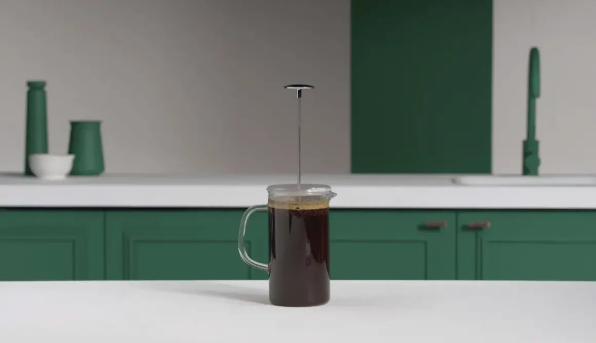En kaffepress med mörkt kaffe redo att pressas. Köksbänk med gröna skåp och en kran i bakgrunden.