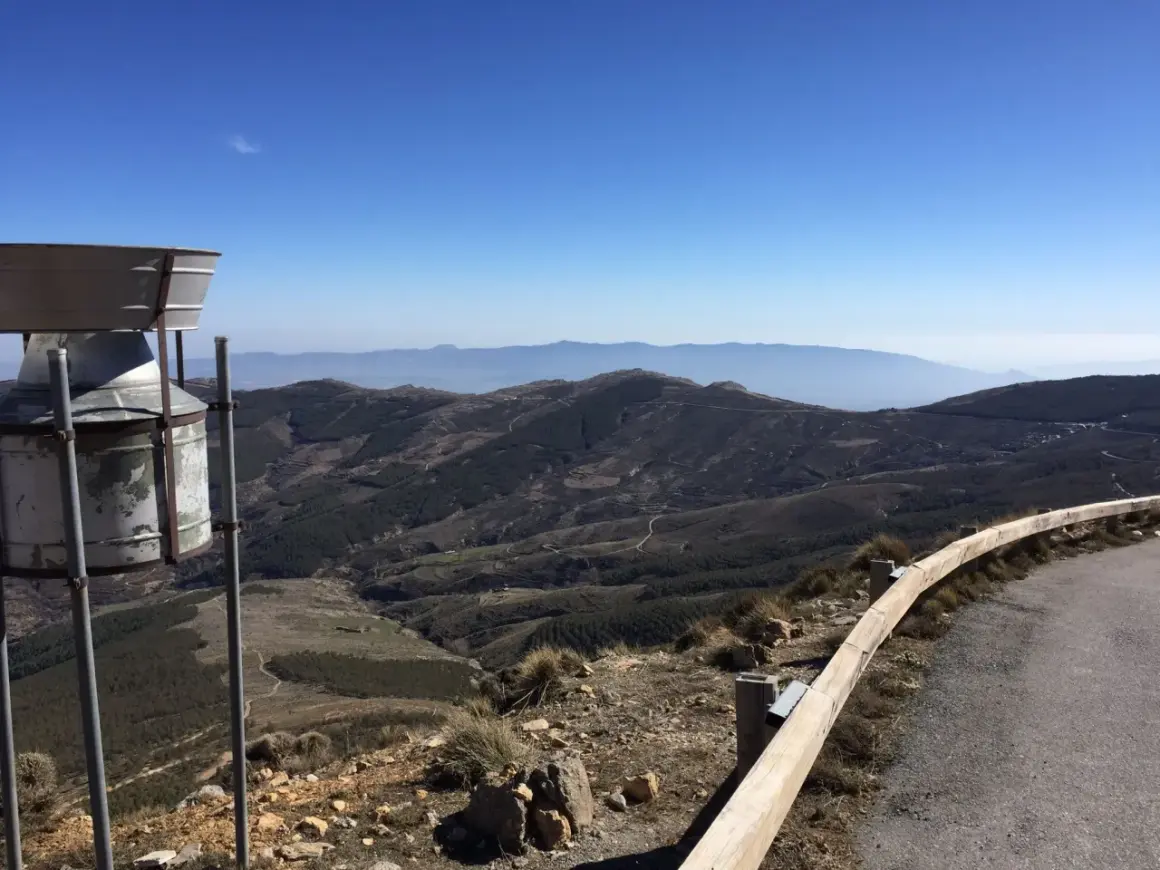 Vista panorámica desde la tetica de bacares, con un pluviómetro en primer plano y un paisaje montañoso y boscoso al fondo bajo un cielo azul.