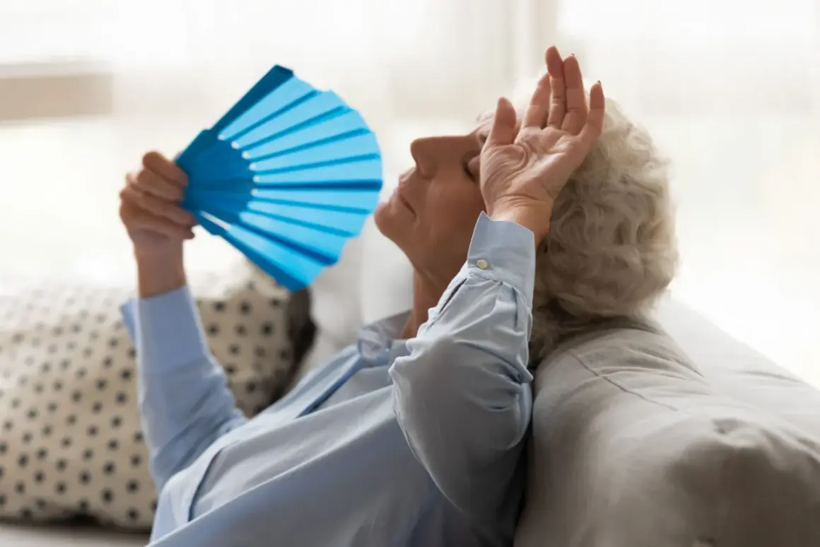 Starsza kobieta zmagająca się z uderzeniami gorąca, wachlująca się niebieskim wachlarzem. Może to być związane z menopauzą, którą leczy fezolinetant.