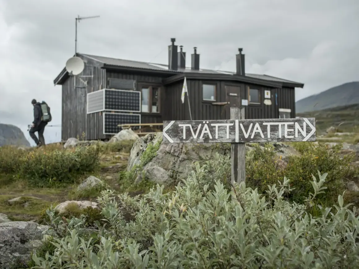 Vandrare närmar sig en av kungsleden stugor. En skylt pekar mot "TVÄTT VATTEN".