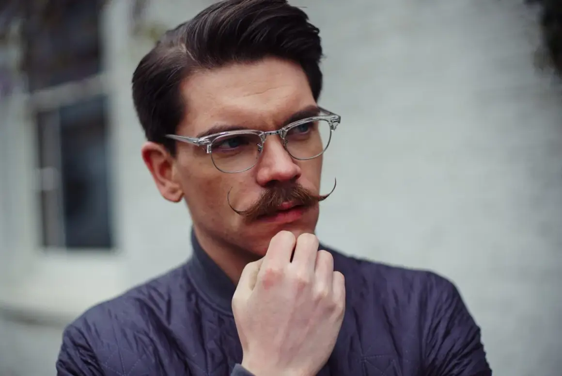 Un homme au visage losange, lunettes fines et moustache élégante, réfléchit.