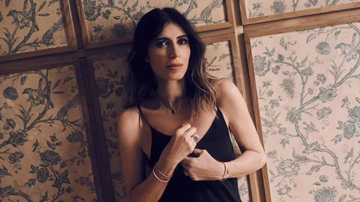 Woman in black slip dress leans against a wall with floral wallpaper.