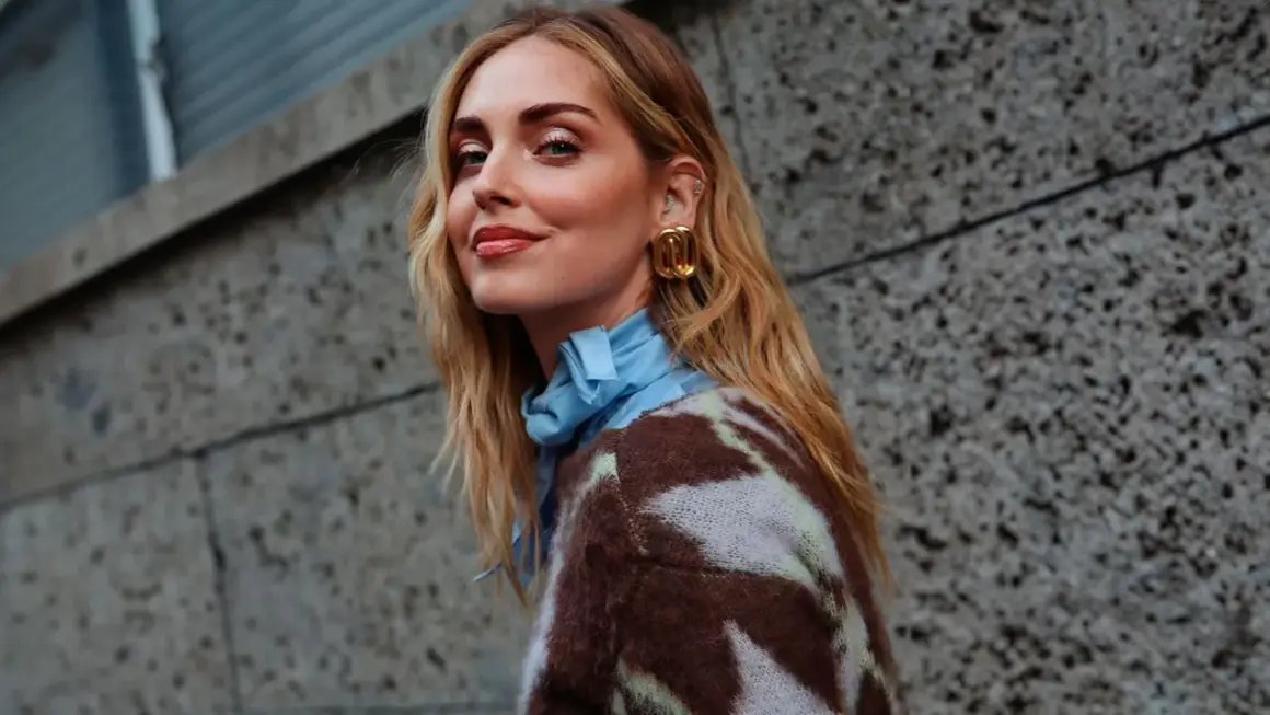 Mujer con media melena rubia, pendientes dorados y jersey estampado, sonriendo ante un muro de hormigón.