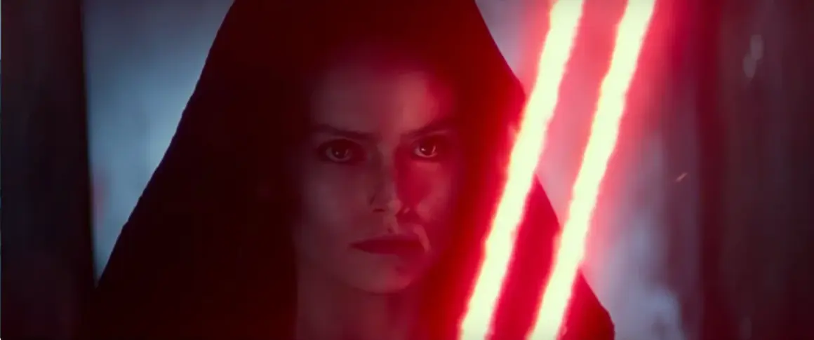 Rey Skywalker w kapturze, z ponurym wyrazem twarzy, trzyma podwójny czerwony miecz świetlny.