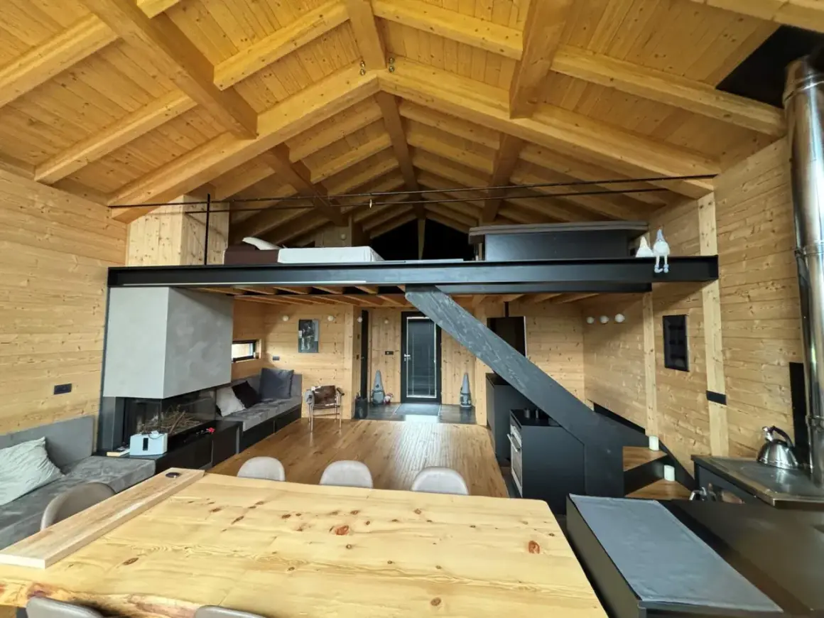 Interno di una baita con splendidi tetti in legno a vista, camino moderno, soppalco e arredi accoglienti.