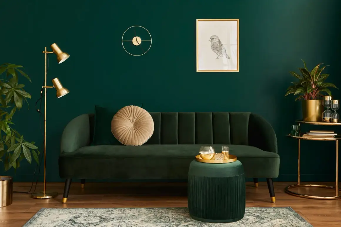 Elegancki salon z butelkową zielenią ściany. Miękka sofa, złota lampa i dekoracje tworzą przytulną atmosferę.