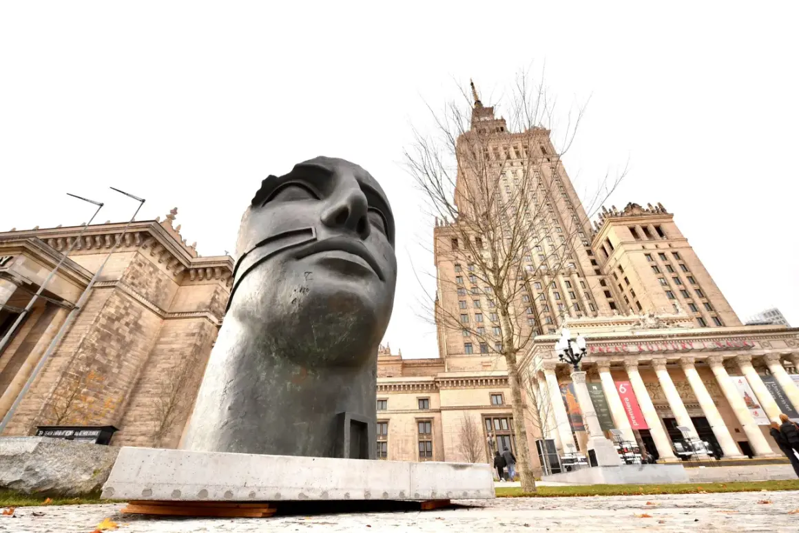 Monumentalna rzeźba głowy na tle Pałacu Kultury w Warszawie.