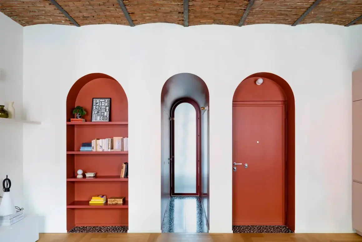 Nicchia nel muro con mensole color terracotta, piena di libri e oggetti decorativi. Tre archi bianchi incorniciano la nicchia, un corridoio e una porta rossa.