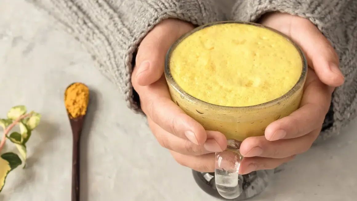 Warme goldene Milch in einer Tasse, gehalten von Händen in einem Strickpullover. Daneben ein Löffel mit Kurkumapulver. Die goldene Milch Wirkung ist bekannt für ihre entzündungshemmenden Eigenschaften.