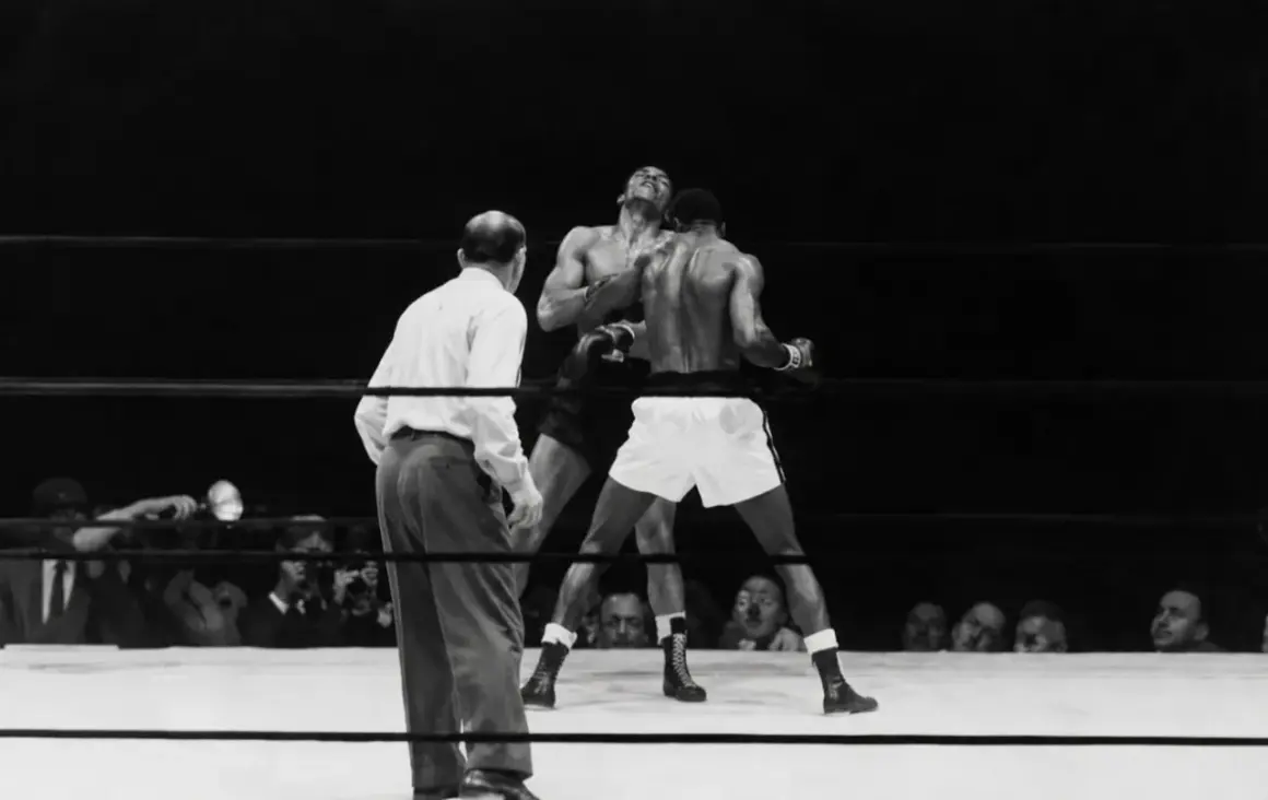 A referee watches as two boxers battle in the ring. One boxer, with his head thrown back, is hit. This intense moment captures the fight for who is the king of boxing.