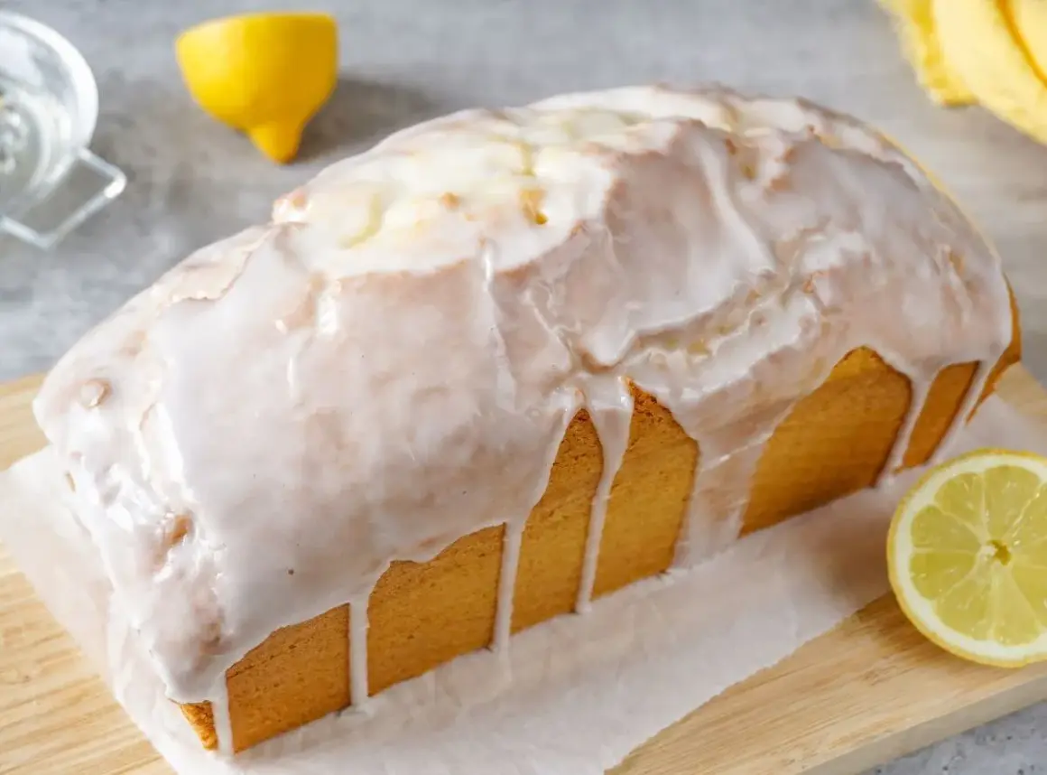 Saftiger zitronenkuchen in Kastenform, überzogen mit süßem Zuckerguss. Ein Zitronenstück liegt daneben.