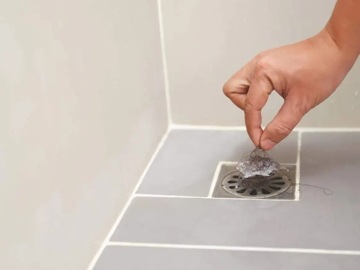 Main d'une personne retirant des cheveux du siphon pour déboucher la douche.