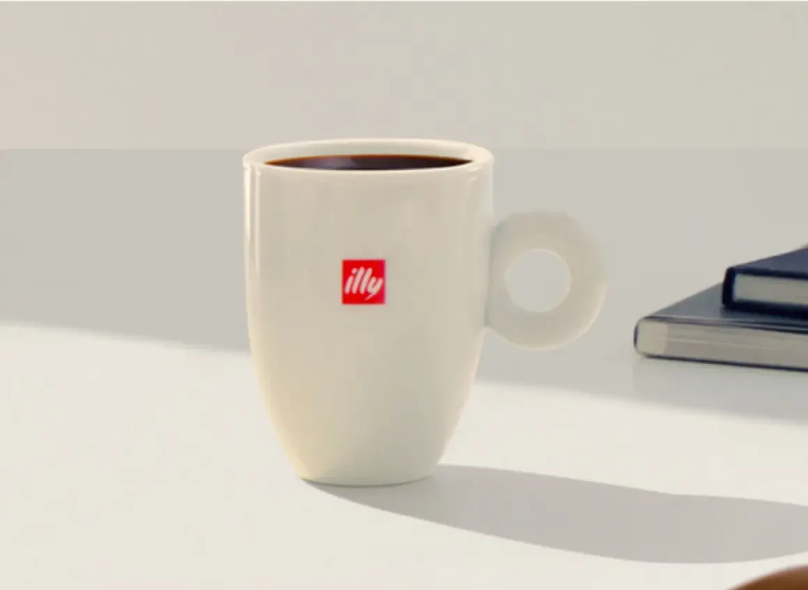 Tazza bianca con logo Illy, piena di caffè americano, pronta per una pausa studio a casa.