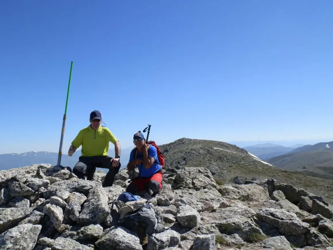 Dos excursionistas celebran en la cima tras una **cuerda larga ruta**. Un poste verde marca el camino.