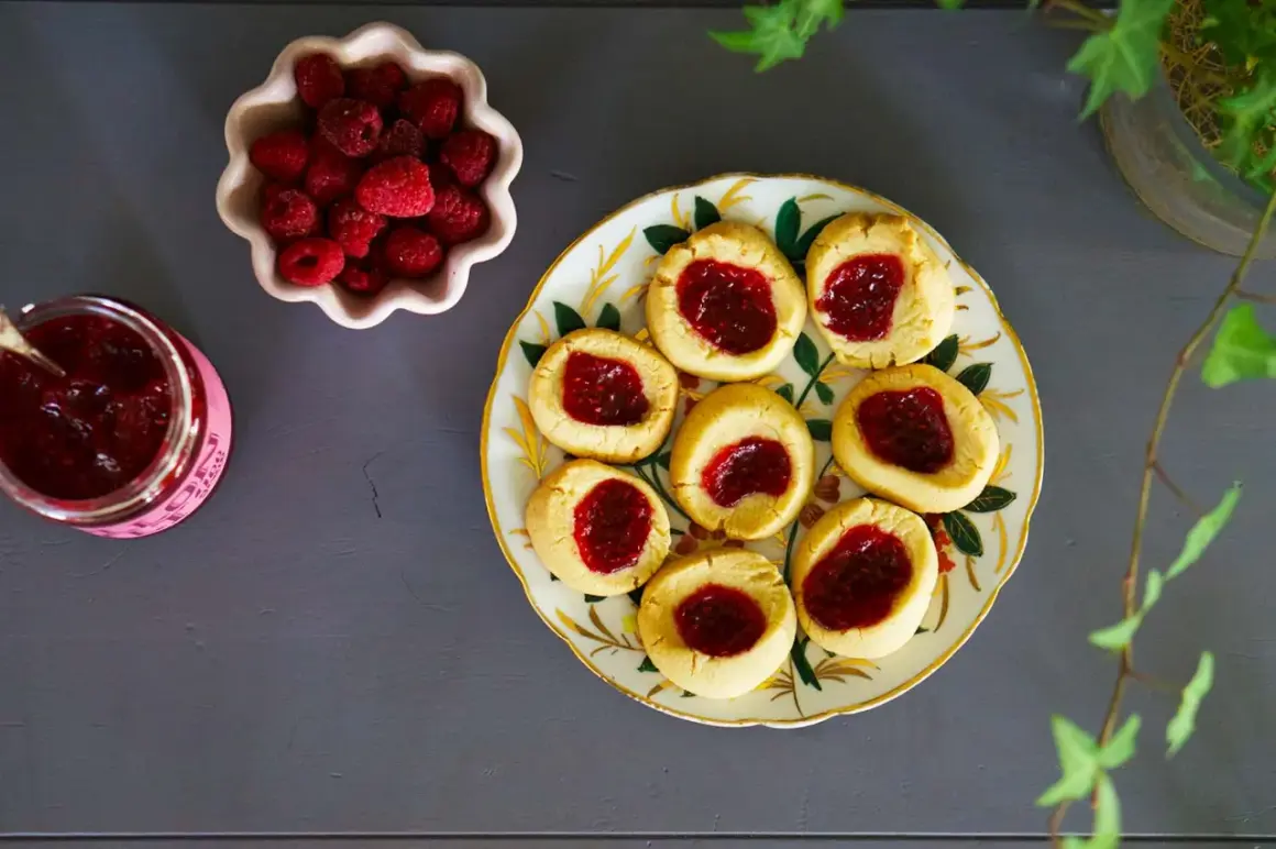 Hallongrottor recept: småkakor med hallonsylt på en vacker tallrik, bredvid en skål med färska hallon och en burk sylt.